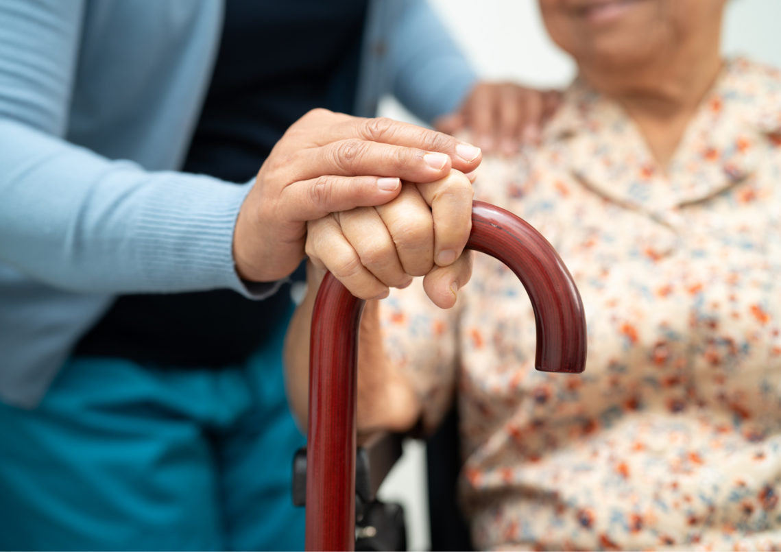 close up of 2 adults. we can't see their faces. focus is on one of heir hands covering the other persons hand, both resting on a walking stick