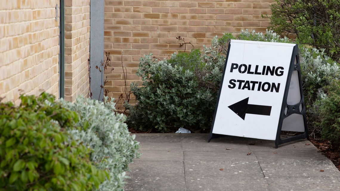 A black and white sign with the words 'Polling Station' and an arrow pointing towards a building entrance along a paved path surrounded by green bushes