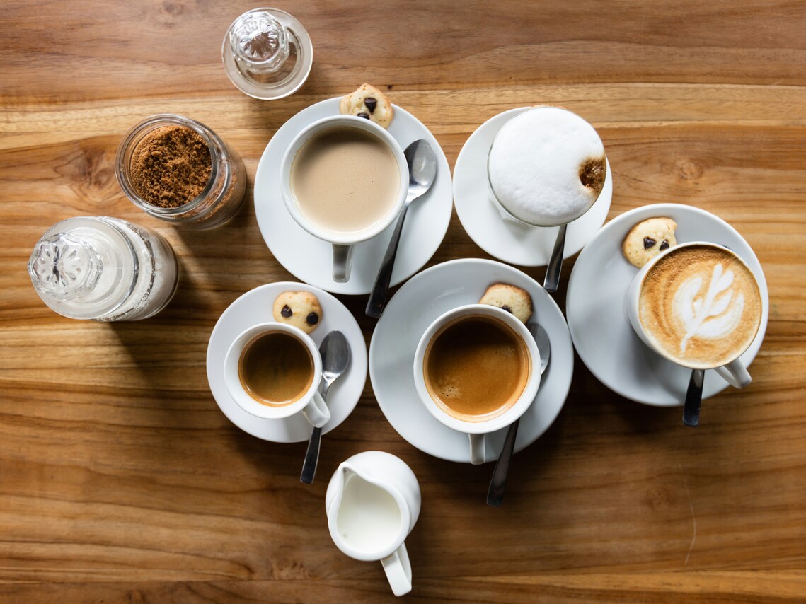 A top down view of cups of coffee in white cups with saucers filled with different types of coffee and teaspoons, small biscuits, a white milk jug, and glass jars of coffee and sugar