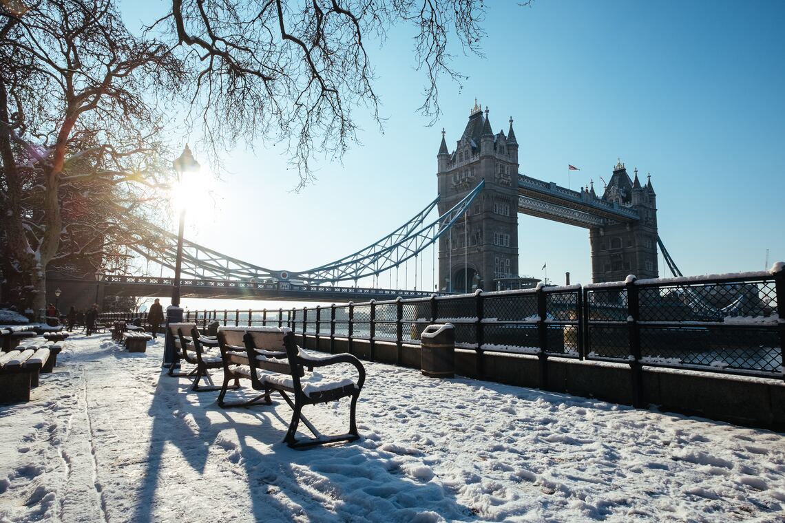 A wooden bench in London covered in snow in the foreground with Tower Bridge in the background